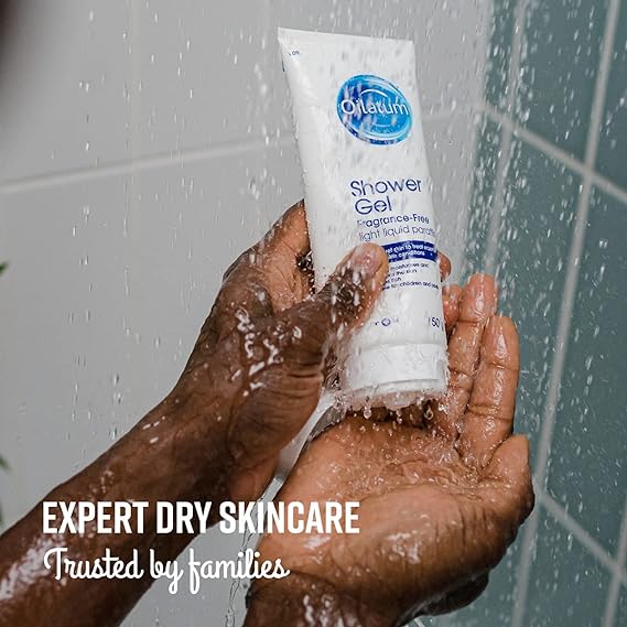 A hand holds Oilatum Shower Gel Fragrance Free (150ml) under a shower stream. Text reads: EXPERT DRY SKINCARE Trusted by families. Tiled bathroom walls emphasize gentle relief for dry, dandruff-prone skin. Brand: Oilatum.