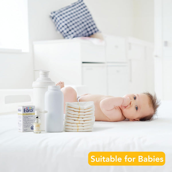 A baby rests on a white bed, surrounded by baby products such as diapers, lotion, and Baby Earol Olive Oil Spray (10ml) by Earol. A yellow label at the bottom right reads Suitable for Babies. The well-lit room has a checkered pillow in the background.