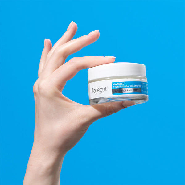 A fair-skinned hand holds a 50ml jar of Fadeout Advanced Brightening Day Cream SPF20 by Fadeout, formulated to reduce dark spots, set against a vivid blue background.