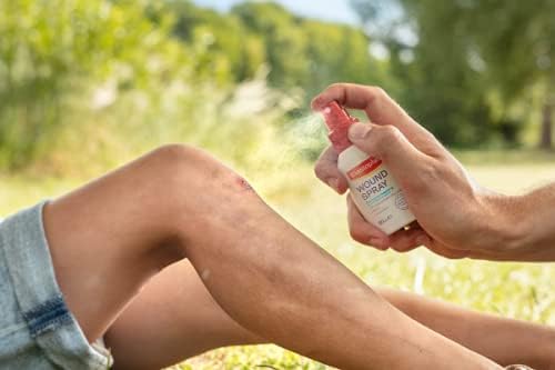 A person gently uses Elastoplast Wound Spray (100ml) on a child’s knee while outdoors. The curious child, sitting on the grass with trees behind, observes this vital step in speeding up the healing process.