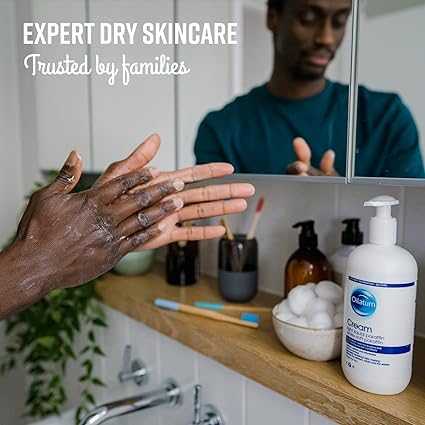 In a bathroom, someone applies lotion to their hands. The shelf features Oilatum Eczema Dry Skin Emollient Cream (500ml) along with toiletries like a toothbrush and cotton balls in a cup. Text reads: EXPERT DRY SKINCARE Trusted by families dealing with eczema.