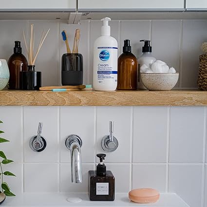 A bathroom shelf displays skincare and hygiene items, including a 500ml bottle of Oilatum Eczema Dry Skin Emollient Cream. Nearby are a soap dispenser, jars with cotton balls, toothbrushes, and a reed diffuser. Below, the sink has silver faucets, a soap bar, and another dispenser on the counter.