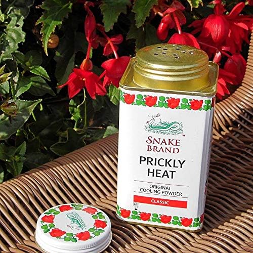 An open container of Snake Brand Prickly Heat Cooling Powder (150g) rests on a wicker surface, providing soothing relief for skin irritation amidst vibrant green leaves and red flowers. The floral-patterned, Classic labeled container is the perfect companion for hot weather comfort.