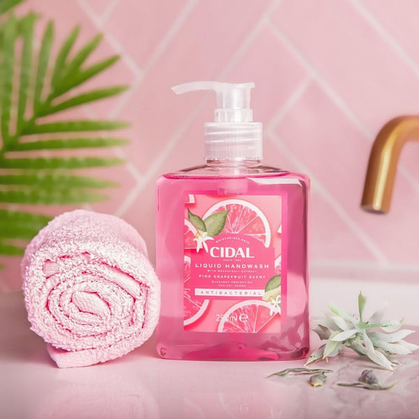 A 250ml bottle of Cidal Pink Antibacterial Liquid Handwash Grapefruit stands beside a pink towel, green plant, and gold faucet on a pink countertop, highlighting hygiene and the fresh grapefruit scent.