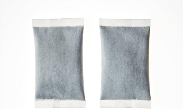 Two Hot Hands desiccant packets rest on a white background. Each gray, rectangular packet, made from thin fabric with sealed edges, resembles hand warmers. Positioned side by side with a small gap, they evoke warmth for chilly weather or outdoor activities.