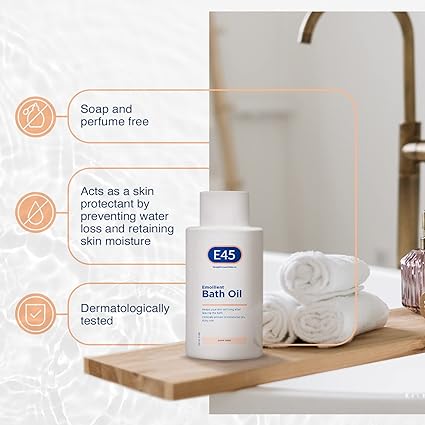 E45 Emollient Bath Oil (500ml) sits on a wooden tray by the sink with rolled white towels. Its soap and perfume-free, retains moisture to protect skin, and is dermatologically tested for dry skin conditions.