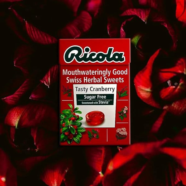 A Ricola Swiss Herb Drops Sugar-Free Cranberry Box (45g) is set among dark red petals, with its Sugar-Free label in white text on a rich red background, sweetened with Stevia for a fresh twist.