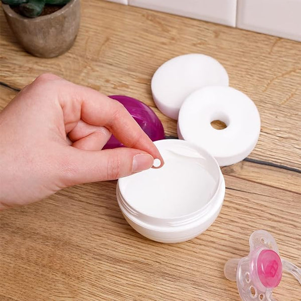 A hand holds a small round Milton Mini Sterilising Tablet (50 Tablets) over an open container on a wooden surface, surrounded by a purple cap, white lid, and pink pacifier—products designed to sterilize baby soothers for enhanced immune system protection.