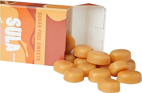 An open box of Sula Sugar Free Sweets - Butterscotch (42g) reveals caramel-colored, naturally flavored candies spilling out. The packaging highlights Sulas orange and white design, showcasing the delightful allure of their butterscotch treats.