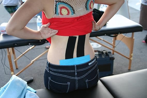 A person, back to the camera, wears a red top and jeans with RockTape Kinesiology 5cm Tape in Electronic Blue applied for injury support on their lower back. The blue tape is visible under the jeans’ elasticity as a massage table waits in the foreground.