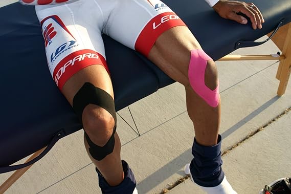 A person in cycling shorts sits on a massage table outdoors on a sunny day with electronic blue RockTape Kinesiology Tape supporting their knees, providing elasticity for optimal recovery.