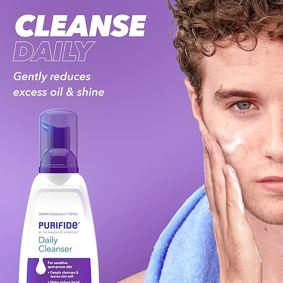 With curly hair, someone applies a white cream to their cheek as part of their skincare routine. Nearby, Galdermas Purifide Daily Cleanser 235ml, designed for sensitive, spot-prone skin, rests against a purple background. The text states: CLEANSE DAILY. Gently reduces excess oil & shine.