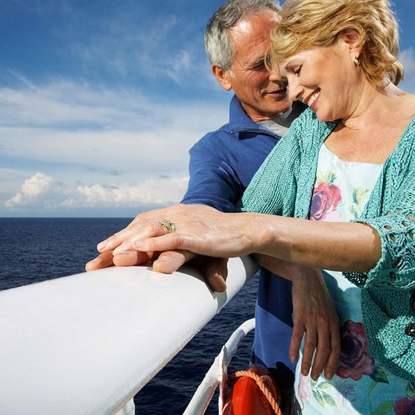 An elderly couple, smiling and holding hands on a ships deck. The woman in a floral dress with a turquoise cardigan, the man in a blue shirt. Although they occasionally face travel sickness, the Sea-Band Nausea Relief Ginger Cap (20) helps them enjoy the blue ocean and clear sky backdrop.