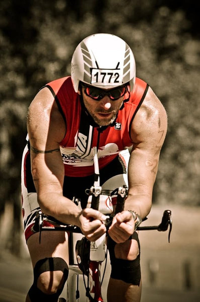 Wearing a red and white triathlon suit and a helmet marked 1772, the cyclist leans into the aero bars of their road bike. RockTapes Electronic Blue Kinesiology Tape supports their muscles, boosting performance. The blurred background captures their rapid movement.