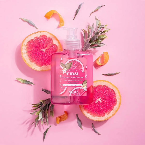 A bottle of Cidal Pink Antibacterial Liquid Handwash Grapefruit (250ml) rests on a pink background, surrounded by grapefruit halves, green leaves, and orange peel pieces for a vibrant hygiene display.