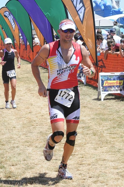 Male athlete, bib 1772, races in a triathlon wearing red and white, with sunglasses and a red cap. He uses knee supports and RockTape Kinesiology 5cm Tape in Electronic Blue for elasticity. Other participants and colorful flags stream by in the background.