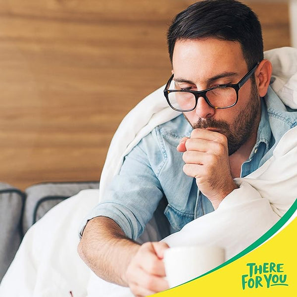 A man with glasses sits on a couch wrapped in a blanket, holding a steaming cup of Lemsip All-in-One in one hand and covering his mouth with the other as he coughs. The words There for You are written in green on a yellow section at the bottom, promising colds and flu relief.