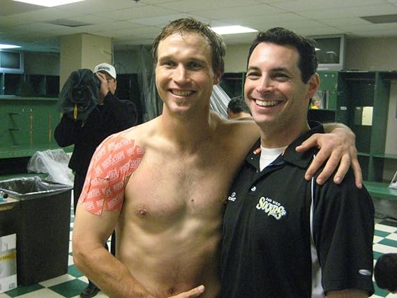 In a locker room, two men smile warmly. The shirtless man on the left proudly displays his resilience with a wrapped shoulder using RockTapes Electronic Blue Kinesiology Tape. The other man sports a black polo. They pose together, showcasing camaraderie and strength amidst injuries.