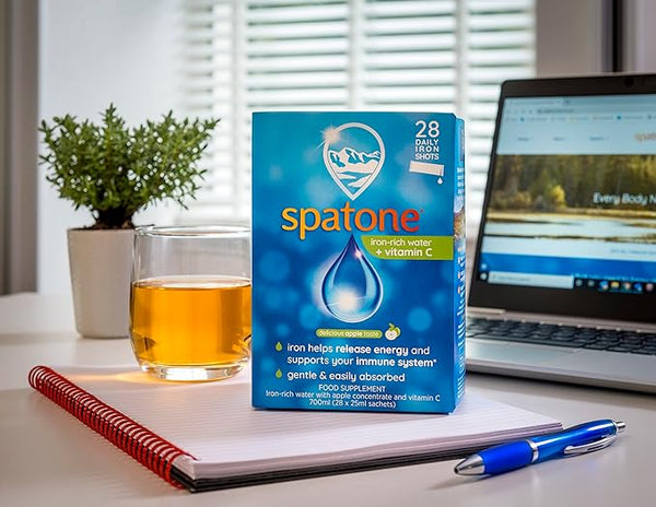 A box of Spatone Apple Daily Iron Shots + Vitamin C (28 Sachets) is on the table next to a glass with the liquid, surrounded by a laptop, notepad, and pen. A leafy plant is in the background as sunlight streams through the window.