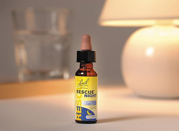 A bottle of Bach Rescue Night Dropper stands on a surface, promising tranquility for sleep with its natural ingredients. The label is yellow and blue, featuring a moon image. A lamp and glass of water in the background enhance the serene atmosphere it creates.