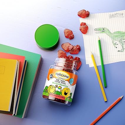 An open jar of Seven Seas Haliborange Mr Men Multi-Vitamin Softies rests on a table with gummies spilling out. Nearby are colored pencils, a green lid, and a dinosaur drawing on lined paper, while books are stacked on the left.