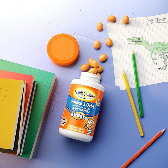 A bottle of Seven Seas Haliborange Kids Omega 3 Chewable Capsules (90) sits among colorful books, pencils, an orange lid, and a childs Raptor dinosaur drawing. These capsules provide vital nutrients for cognitive and digestive health.