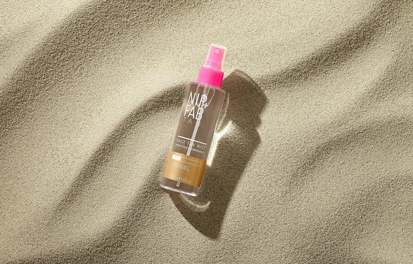 The Nip+Fab Faux Tan Mist Caramel (150ml) bottle, with pink and white labeling and a pink cap, sits on textured sand. It offers a streak-free self-tanning solution in the Caramel shade while shadows play across the surface.