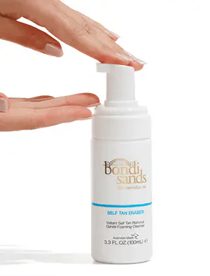 A hand dispenses Bondi Sands Self Tan Eraser (100ml) foam from a white bottle with a blue stripe, highlighting the ideal Bondi Sands solution to remove self-tan and correct tanning mistakes against a simple white background.