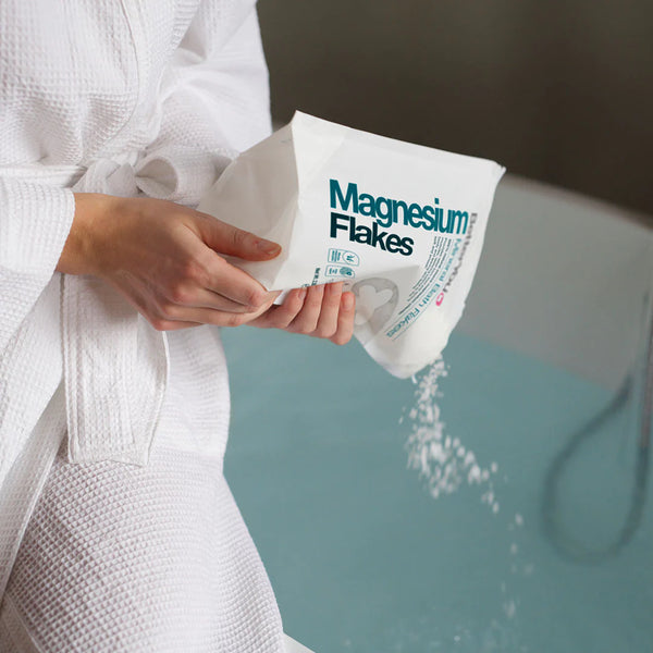 Dressed in a white robe, someone pours BetterYou Magnesium Flakes - Foot & Body Soak (1kg) into a water-filled bathtub for a calming soak.