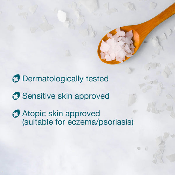 A wooden spoon filled with BetterYou Magnesium Flakes - Foot & Body Soak (1kg) coarse crystals is on a light surface. Text on the left reads: Dermatologically tested, Sensitive and atopic skin approved. Scattered salt crystals lie nearby.