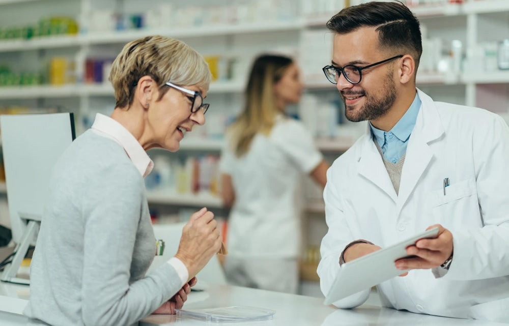 A pharmacist with glasses shows a tablet to an older woman in a pharmacy. Shelves filled with medicine are visible in the background, and another staff member is working nearby.