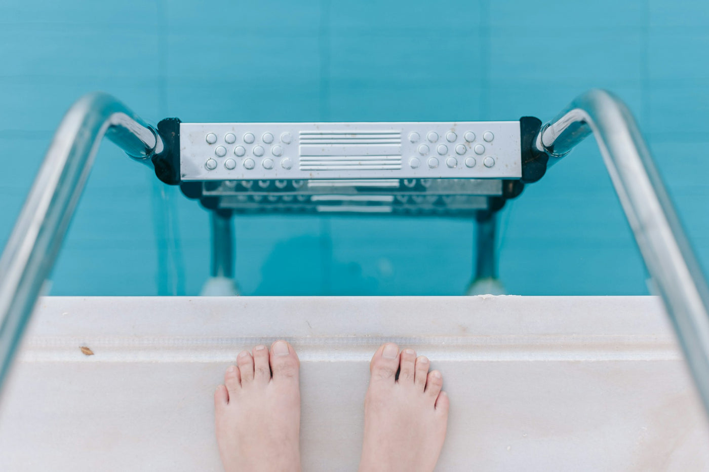 A pair of bare feet stands at the edge of a swimming pool, facing a silver pool ladder that descends into clear, blue water. The scene captures the moment before entering the pool.