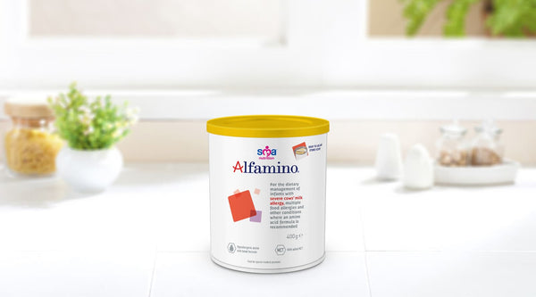 A tin of Sma Nutrition Alfamino (400g), a hypoallergenic formula, rests on a white countertop. In the blurred background, vibrant green plants enhance the scene next to a white sugar bowl as sunlight pours in through the kitchen window.