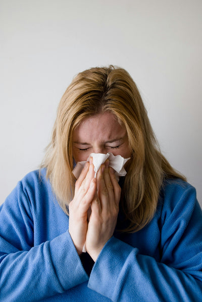 A woman with blond hair is wearing a blue robe and appears to be sneezing or blowing her nose into a tissue. Her eyes are closed, and her expression suggests discomfort. The background is plain and light-colored.