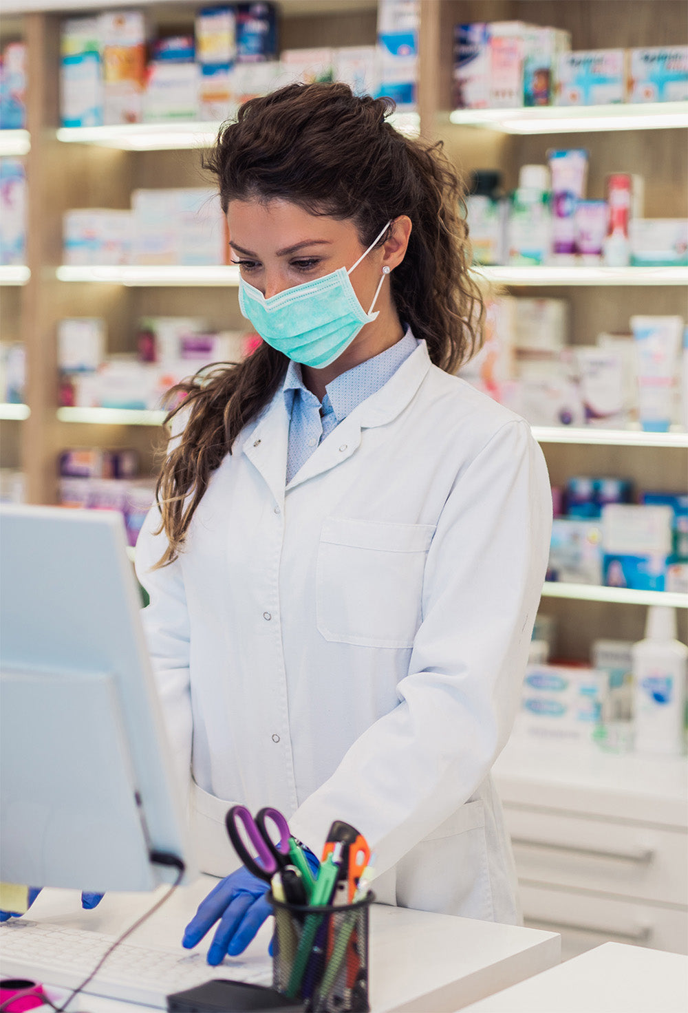 A pharmacist wearing a white coat and a face mask is working on a computer. Behind her are shelves filled with various medicine boxes and products.