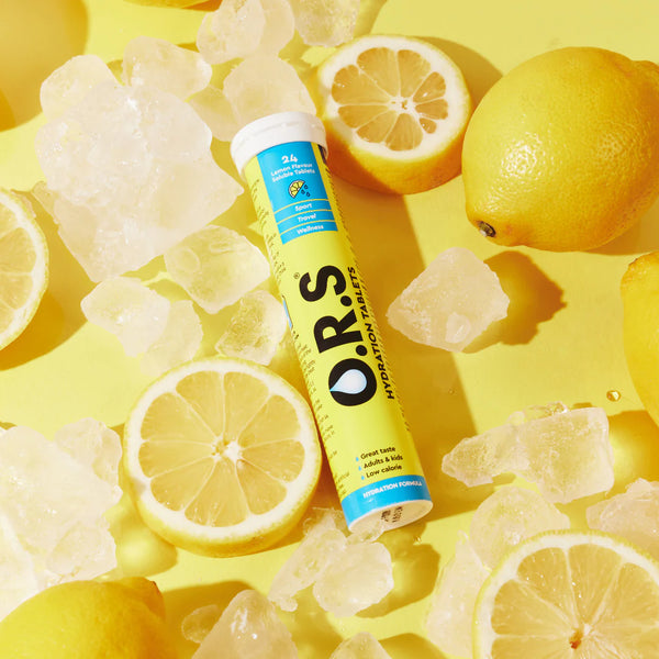 A tube of ORS Hydration Tablets Lemon (24 Tablets) by ORS rests on a yellow surface, surrounded by ice cubes and fresh lemons. The yellow and blue packaging emphasizes effective electrolyte hydration.