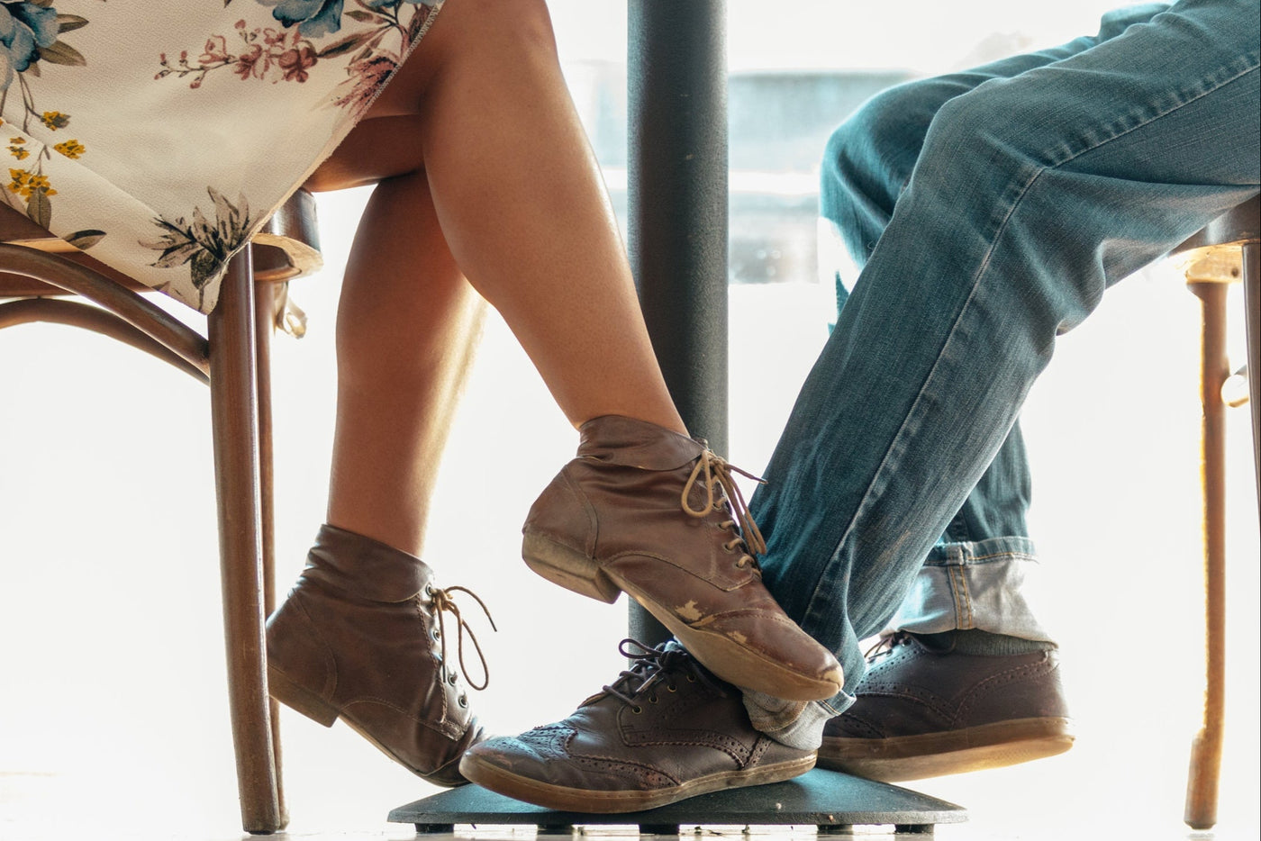 A woman wearing a floral dress and brown shoes playfully intertwines her legs with a man in jeans and brown shoes under a table, suggesting a lighthearted, intimate interaction.