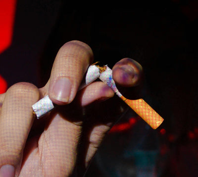 A hand is shown breaking a cigarette in half against a dark background. The cigarette is held between the thumb and forefinger, with tobacco partially spilling out. Red light illuminates part of the scene.