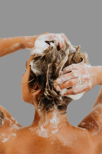 Person washing their hair with shampoo, their back facing the camera. Hands are lathering the hair with white foam against a plain gray background.