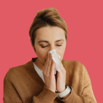 A person with light brown hair is wearing a brown sweater and holding a tissue to their nose with closed eyes, appearing to sneeze or blow their nose. The background is a solid pink color.