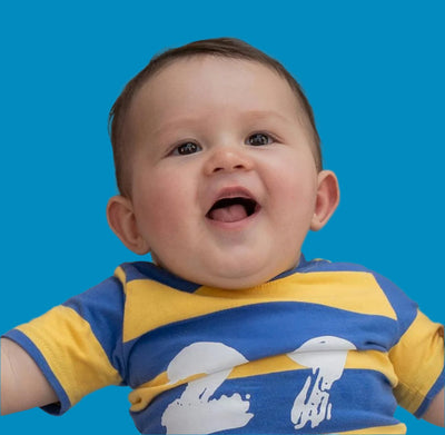 A smiling baby with short brown hair and blue eyes is wearing a blue and yellow striped shirt with a playful white pattern. The background is a solid bright blue.