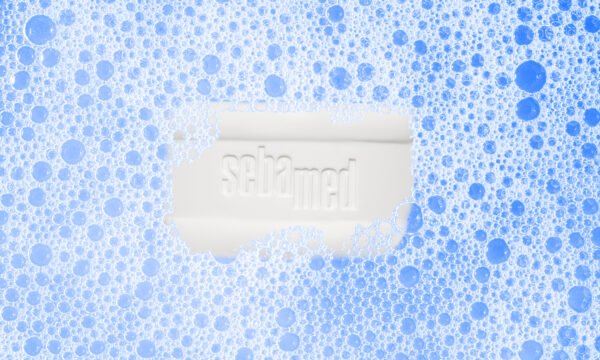 A Sebamed Cleansing Bar 100g for Eczema-Prone Skin Treatment, partially enveloped in blue and white foam bubbles, is centered against a calming blue background—perfect for deep pore cleansing.